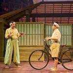 Tatsuo (Dai Tabuchi) wearing a khaki suit and sitting on a hospital bed with Yasuko (Phyllis Ho) and pointing.